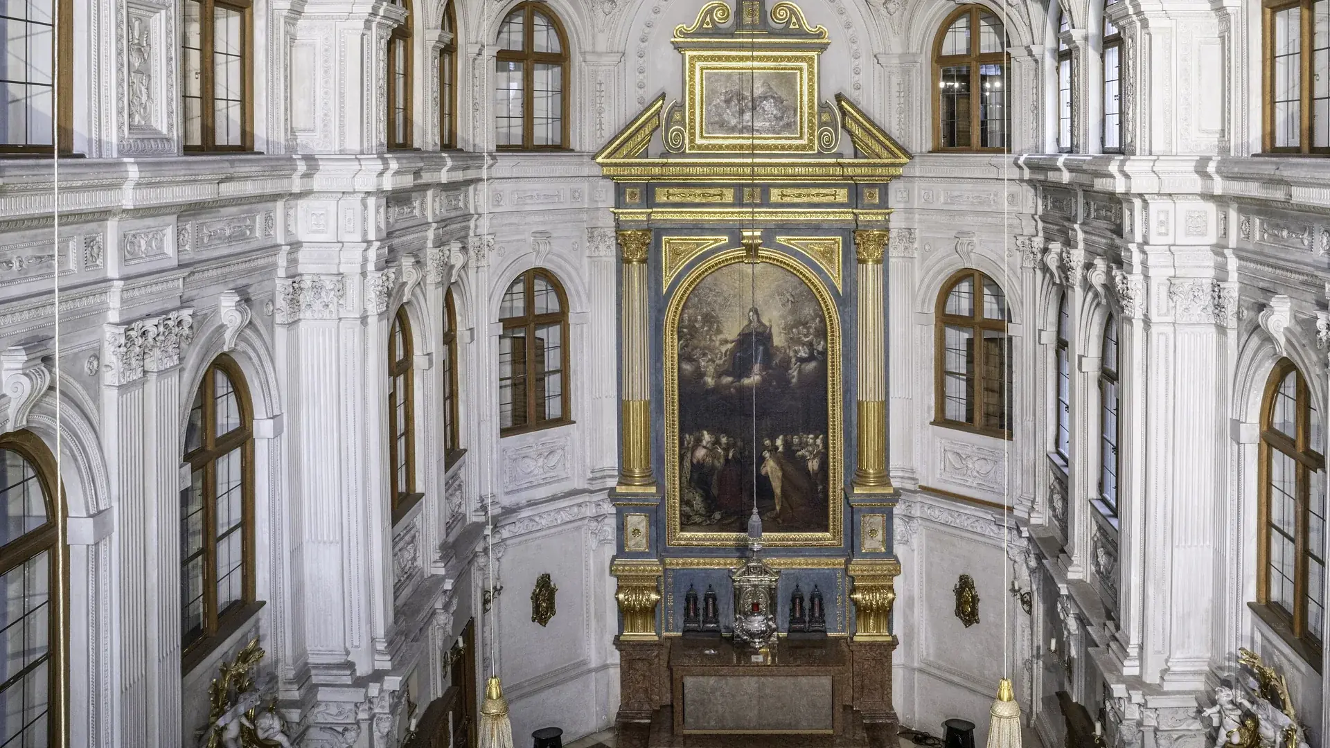 Looking down into the court chapel of the Munich Residenz: white stucco walls, tall arched windows with brown frames, and a gilded altar frame around a large painting of the Virgin in glory.